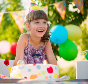 Little girl laughing and sitting in front of a birthday cake while wearing a birthday hat, with balloons in background